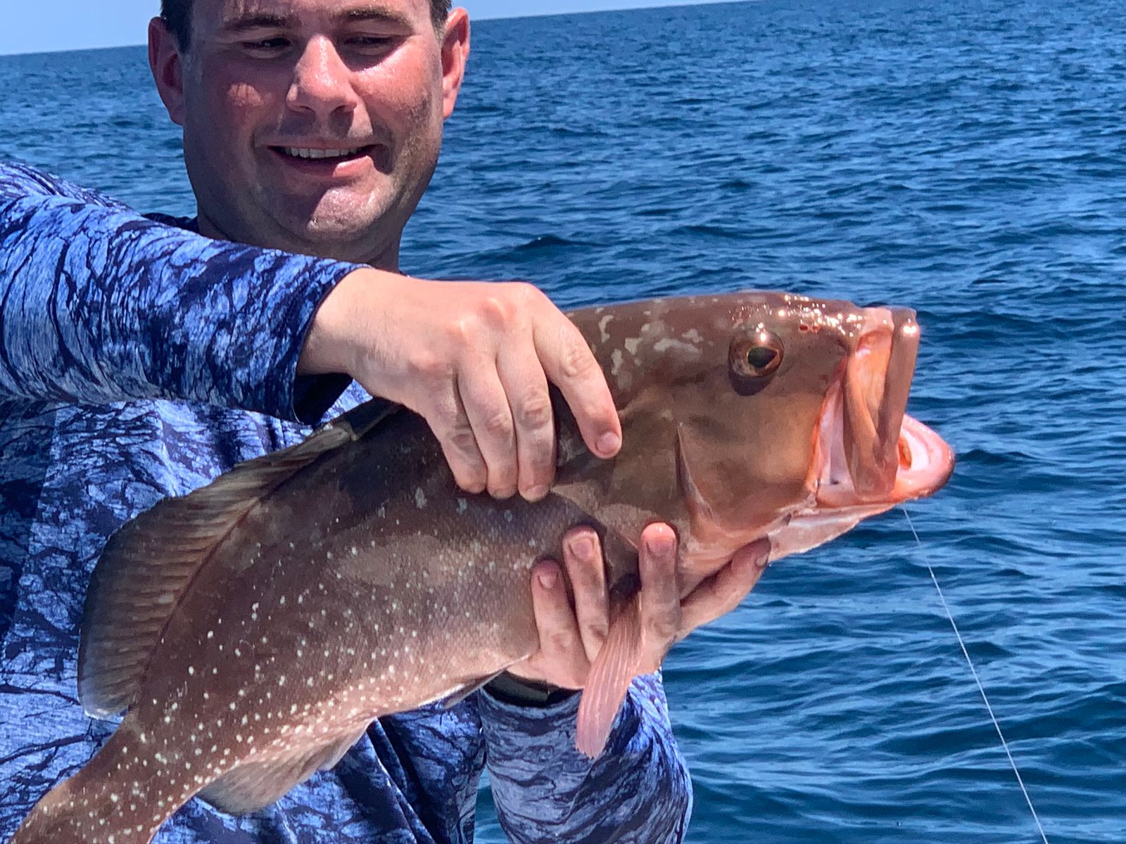 A smiling man holds up a red grouper close to the camera with the open ocean behind him.