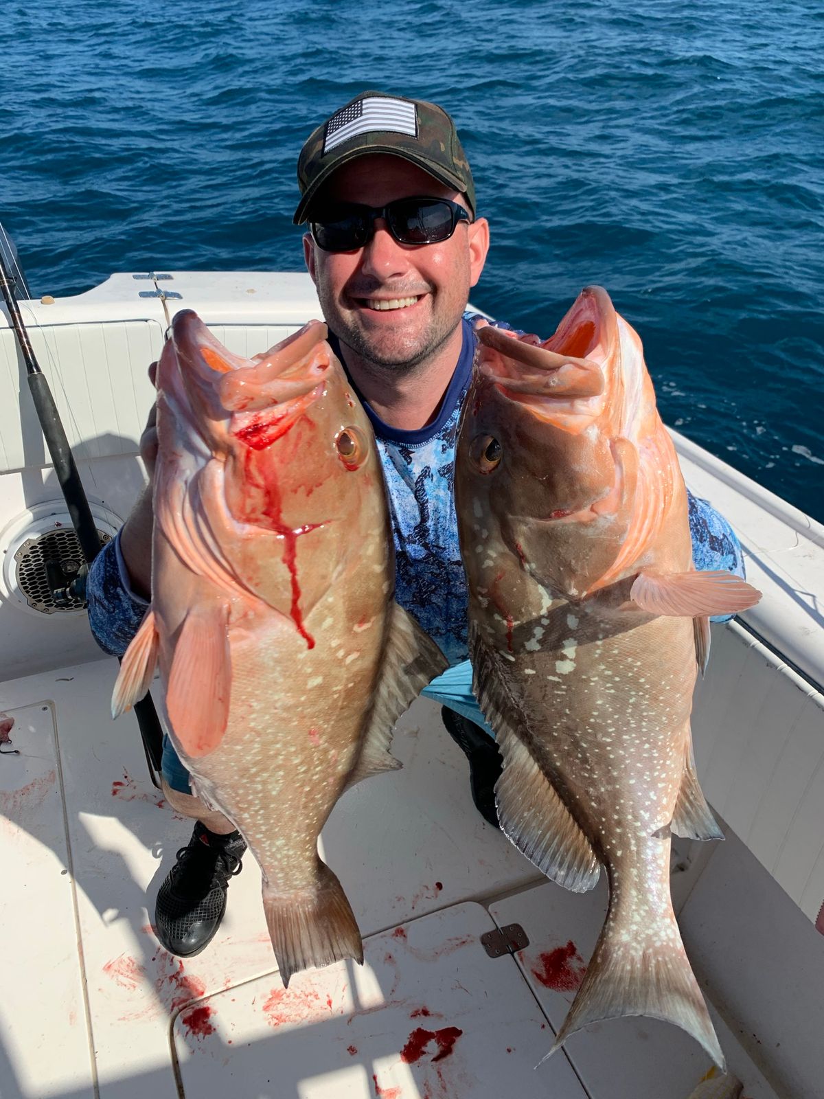 A smiling man holds up two grouper on a boat deck with some blood visible near his feet.