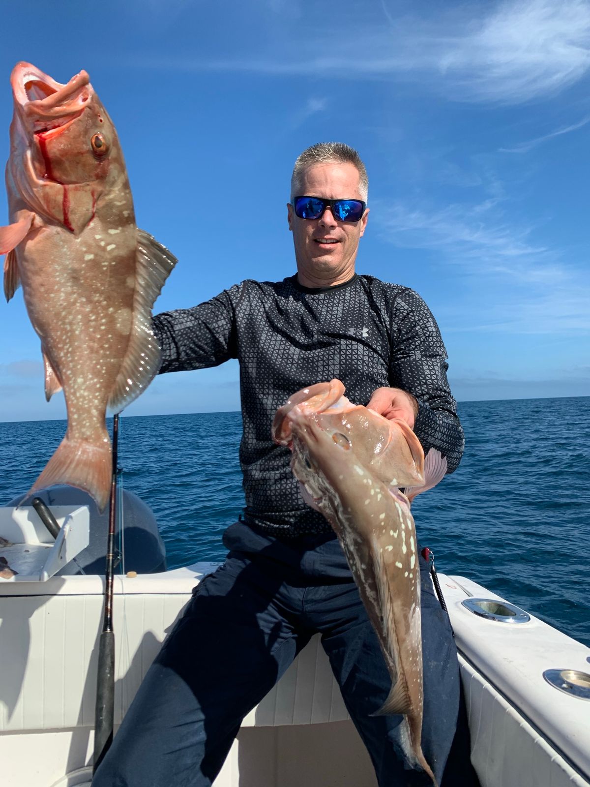 A smiling man holds up a grouper in one hand and another fish in the other on a boat with clear blue sky and water.