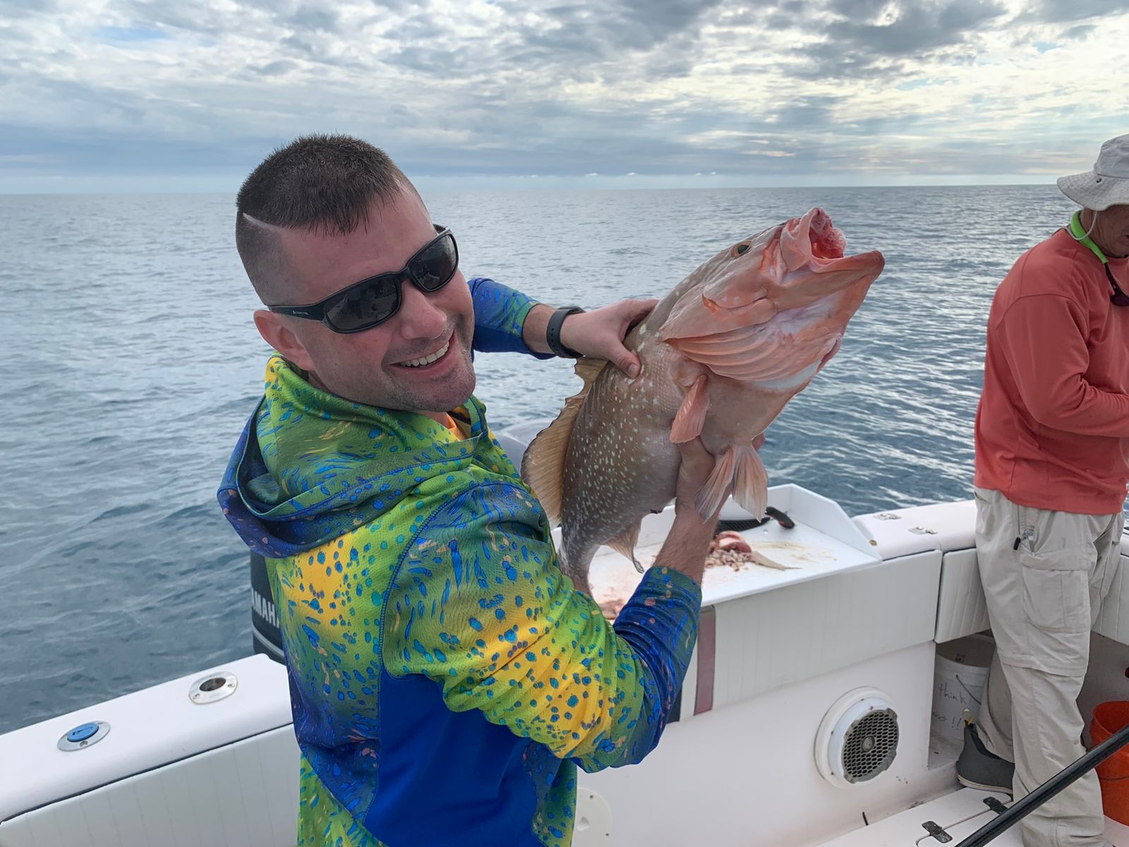 A smiling man holds up a grouper on a boat under a cloudy sky with another angler visible nearby.