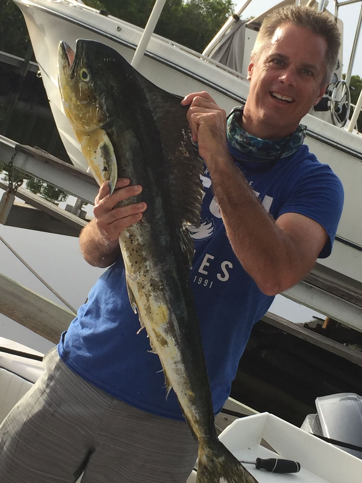 A smiling man holds up a mahi mahi fish at a marina with boats behind him.