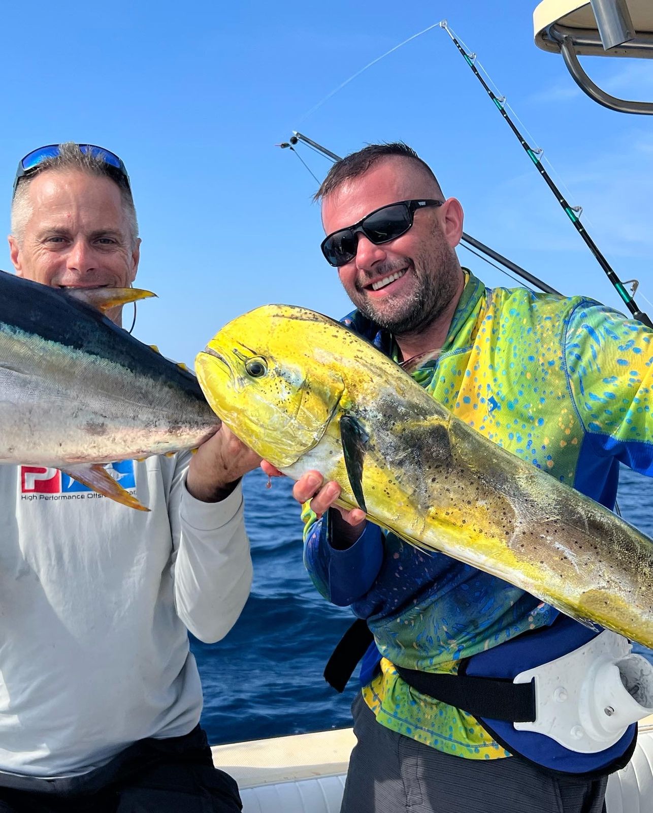 Two smiling men on a boat each hold up a large fish, one yellow mahi mahi and one silver tuna.