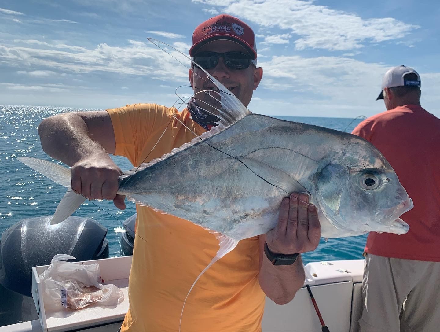A man in an orange shirt holds a large silver fish on a boat with the ocean behind him.