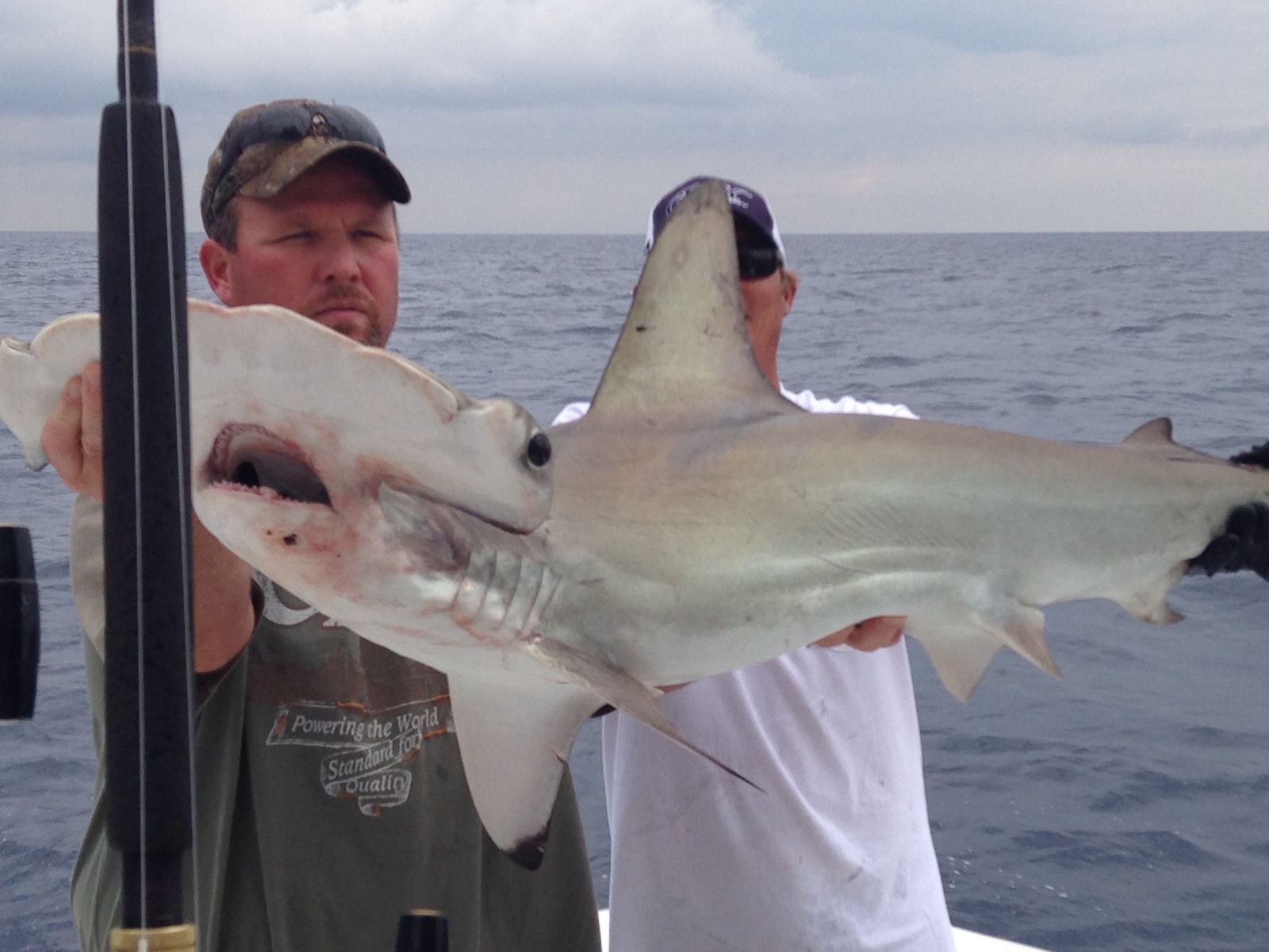 Two men hold up a hammerhead shark on a boat with the ocean and cloudy sky behind them.
