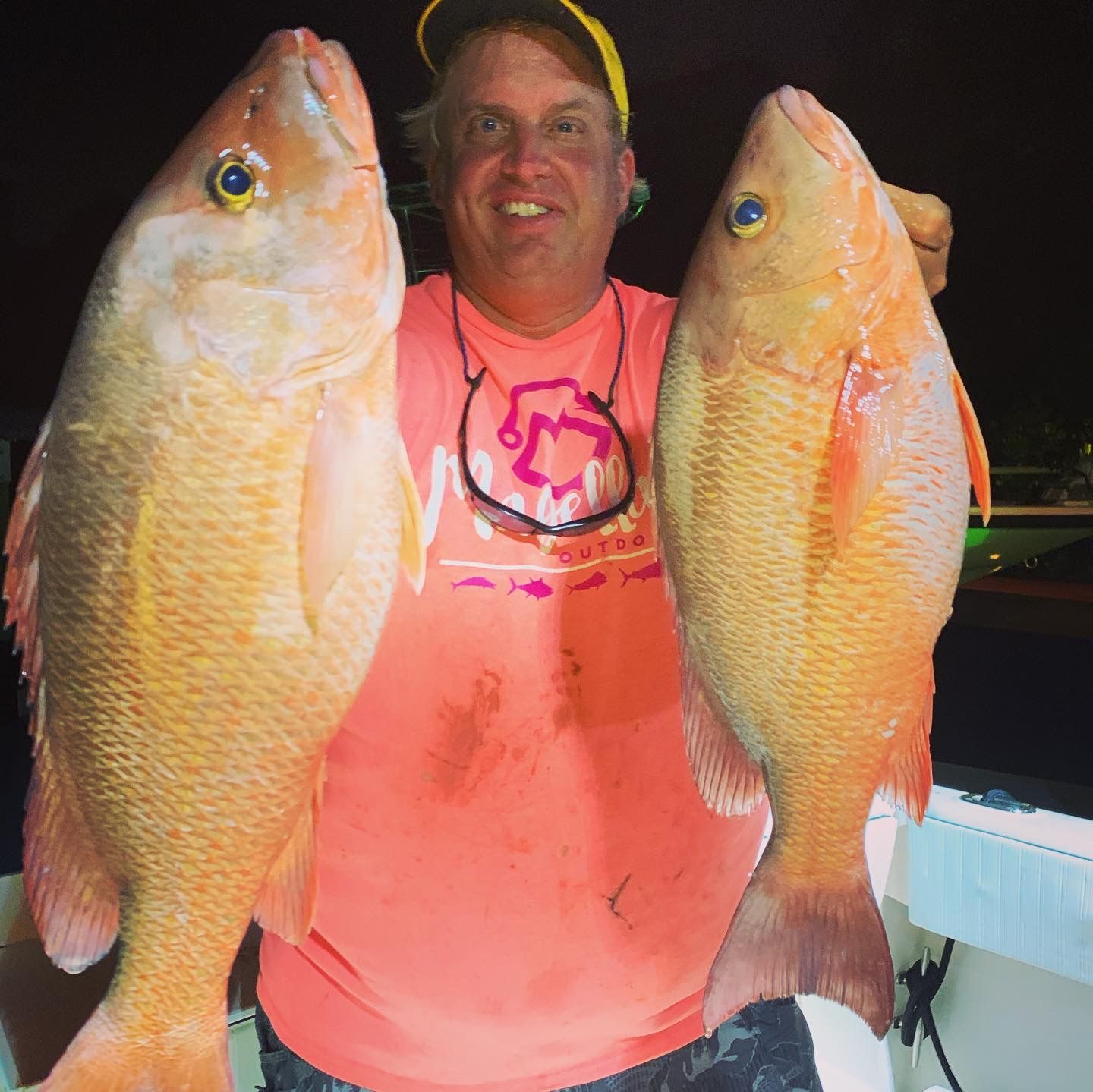 A man holds up two orange snapper fish on a boat deck at night.