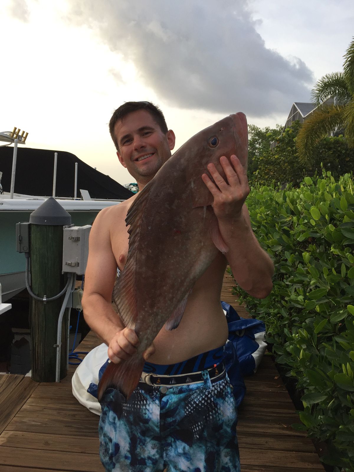 A smiling shirtless man holds up a mutton snapper on a dock at dusk with a boat and greenery behind him.