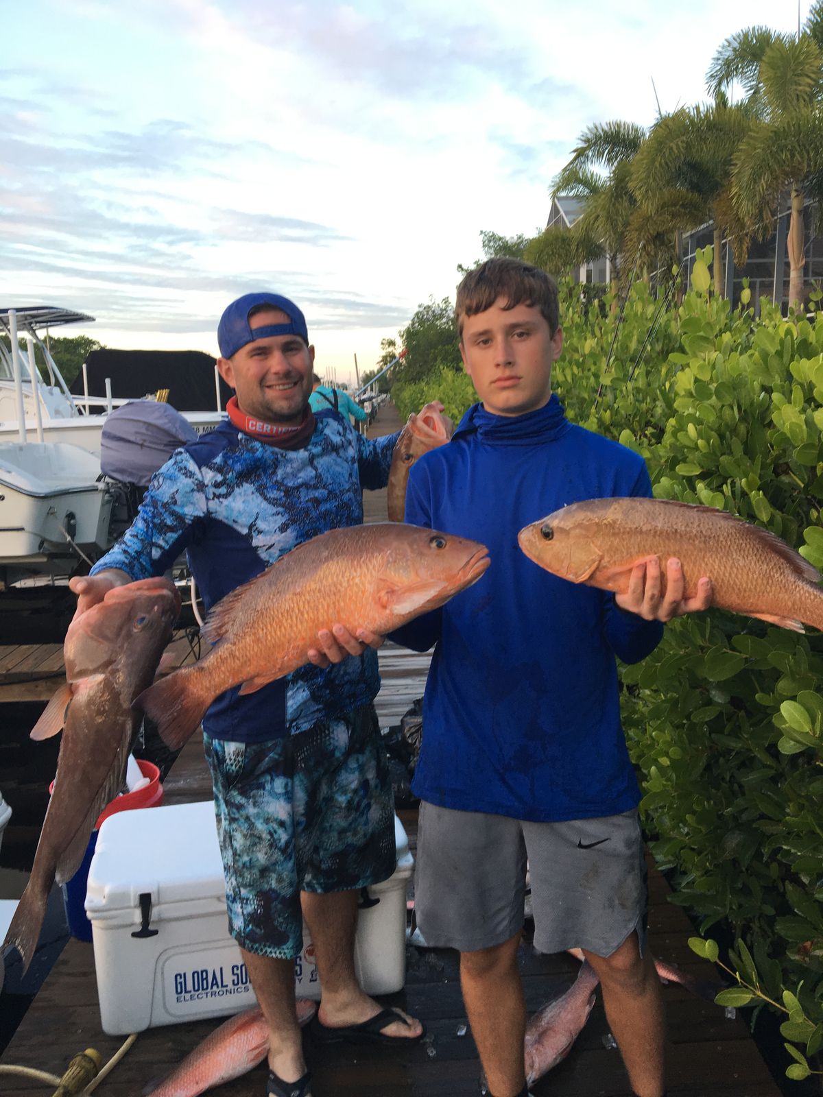 A man and a teenage boy each hold up multiple fish on a dock at dusk with mangroves and boats behind them.