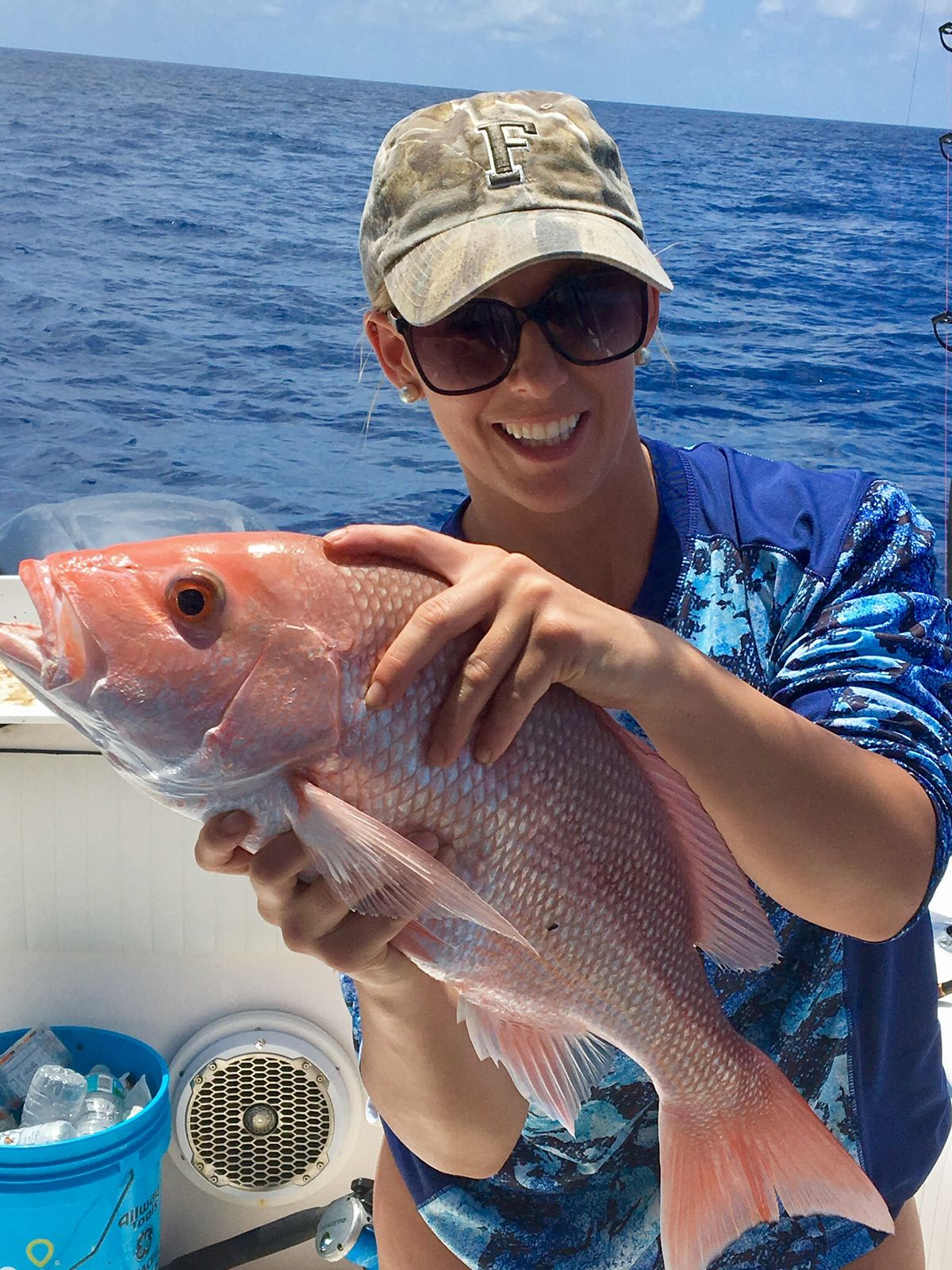 A close-up of a smiling woman holding a red snapper with the ocean behind her.