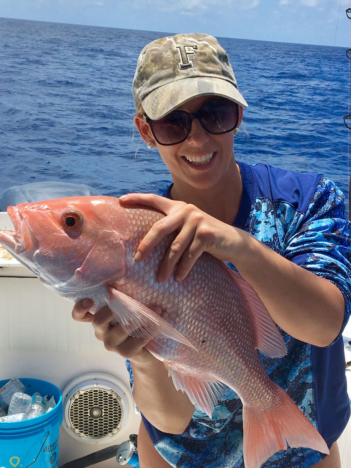 A smiling woman in sunglasses holds up a red snapper on a boat with the ocean behind her.
