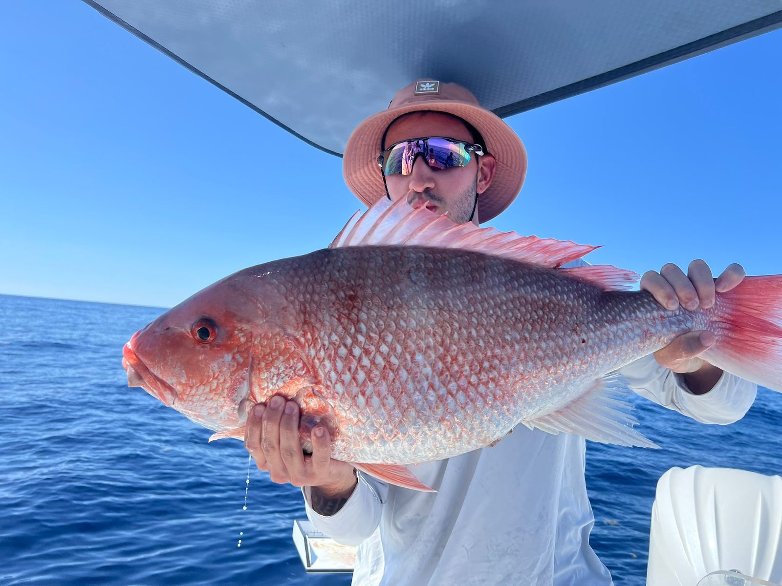 A man in a hat and sunglasses holds a large red snapper close to his face under a boat's shade top with clear sky and water behind him.