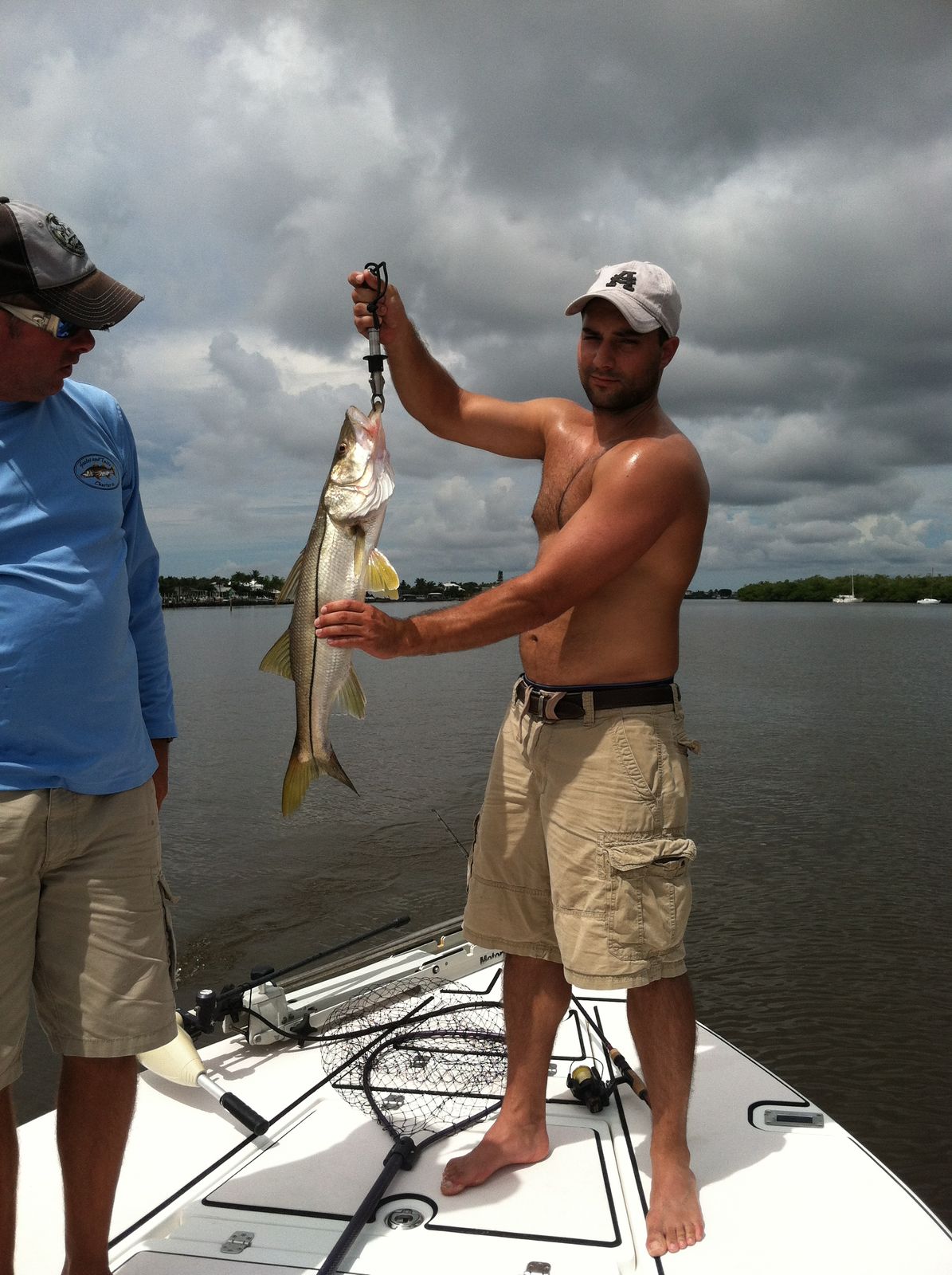 A shirtless man holds up a snook fish on a boat with a cloudy sky and shoreline in the background.